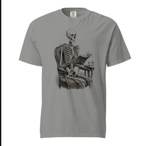 May include: A grey short-sleeve t-shirt featuring a black and white graphic print of a human skeleton sitting in an ornate armchair, reading an open book, with a small side table holding a teacup positioned next to the chair.