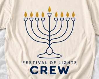 May include: A cream-coloured t-shirt featuring a dark blue line-art graphic of a nine-branched menorah with gold-coloured flames. Below the menorah, the text reads "FESTIVAL OF LIGHTS" in small letters and "CREW" in large, bold blue letters.