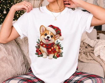 May include: A white short-sleeve t-shirt featuring a graphic of a Corgi dog wearing a red and green plaid Santa hat and a matching scarf. The dog is surrounded by pine branches, red ornaments, pinecones, and glowing yellow string lights.