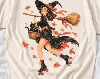 May include: A cream-colored t-shirt featuring a graphic of a witch in a black dress and hat, wearing black roller skates. The witch carries a broom and a basket with a black cat, surrounded by falling orange autumn leaves and red ribbons.