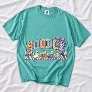 Six poodles in Halloween costumes walking under the arched word BOODLE, lavender and orange polka-dot varsity style