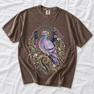 Painted pigeon wearing a daisy crown and glowing halo standing on a fern frond, whimsical cottagecore bird t-shirt art