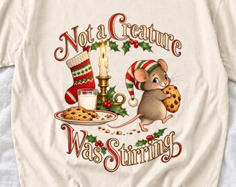 May include: A cream-colored t-shirt featuring a graphic of a mouse wearing a striped nightcap and holding a chocolate chip cookie. The design includes a lit candle, a stocking, a glass of milk, and the text Not a Creature Was Stirring.