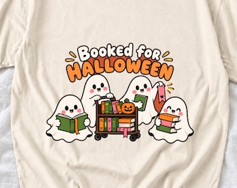 Puede incluir: Una camiseta de color crema con un gráfico de tres fantasmas blancos con caras sonrientes. Los fantasmas interactúan con un carrito de biblioteca marrón lleno de libros coloridos y una pequeña calabaza. El texto dice Booked for HALLOWEEN.