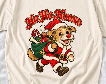 May include: A cream-colored t-shirt featuring a graphic of a dog dressed as Santa Claus carrying a green sack filled with bones and candy canes. The text "HO HO HOUND" is printed in red with a gold outline above the dog, who wears a red hat and suit.