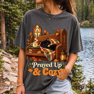 Black cat curled on a church pew with blanket and cocoa among autumn leaves, text "Prayed Up & Cozy", retro illustration