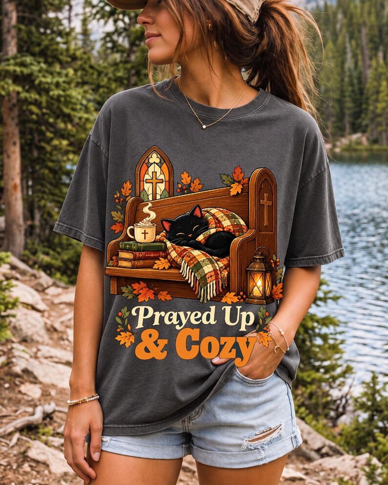 Black cat curled on a church pew with blanket and cocoa among autumn leaves, text "Prayed Up & Cozy", retro illustration
