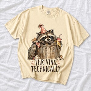 Raccoon in a paper party hat peeking out of a trash can with the text "Thriving, Technically", sepia watercolor sketch