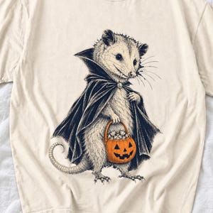 May include: A cream-colored t-shirt featuring a black and white illustration of an opossum wearing a dark cape. The opossum has visible fangs and is holding a small orange jack-o'-lantern bucket filled with white candy pieces.
