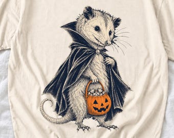 May include: A cream-colored t-shirt featuring a black and white illustration of an opossum wearing a dark cape. The opossum has visible fangs and is holding a small orange jack-o'-lantern bucket filled with white candy pieces.