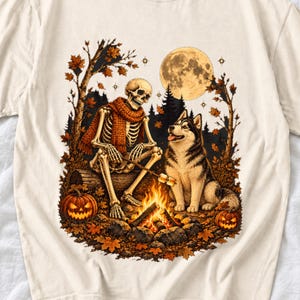 May include: A cream-colored t-shirt featuring a graphic of a skeleton wearing an orange scarf sitting on a log by a campfire with a husky dog. The scene includes a full moon, autumn trees with falling leaves, and two jack-o'-lanterns.