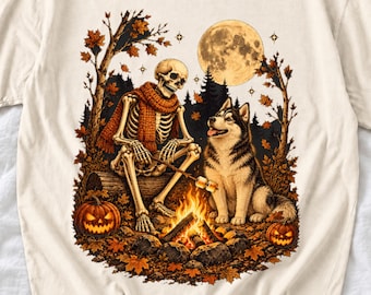 May include: A cream-colored t-shirt featuring a graphic of a skeleton wearing an orange scarf sitting on a log by a campfire with a husky dog. The scene includes a full moon, autumn trees with falling leaves, and two jack-o'-lanterns.
