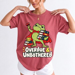 Grumpy cartoon frog librarian holding a stamp and a stack of books with the text Overdue & Unbothered