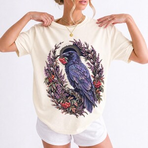 Raven holding a rowan berry inside an oval wreath of heather and catkins, painted cottagecore bird illustration