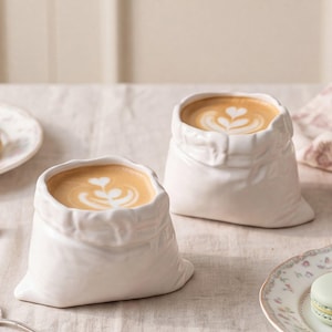 Puede incluir: Dos tazas de cerámica blanca con forma de pequeños sacos de tela plegados, cada una llena de un café con leche con arte en espuma en forma de corazón. Las tazas tienen una apariencia texturizada y arrugada que imita un saco atado.