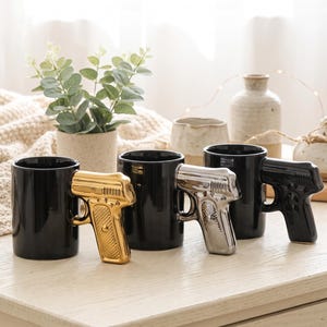 Puede incluir: Tres tazas de cerámica negra, cada una con un asa en forma de pistola. Un asa tiene un acabado en oro metálico, otra en plata metálica y la tercera en negro sólido, a juego con el cuerpo de la taza. Las tazas son de forma cilíndrica.