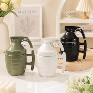 Puede incluir: Tres tazas de cerámica con forma de granada de mano, cada una con un patrón de rejilla texturizado, una palanca superior y una anilla de metal. Las tazas son de color verde oliva, blanco y negro, e incluyen un asa lateral para sujetarlas.