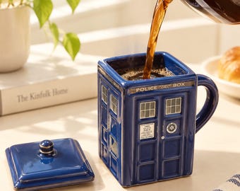 Pode incluir: Uma caneca quadrada de cerâmica azul em forma de cabine de polícia com tampa a condizer. A caneca apresenta janelas brancas, uma pega e o texto POLICE CALL BOX e FREE FOR USE OF PUBLIC PULL TO OPEN. Café escuro está a ser vertido.