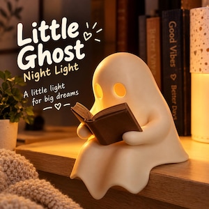 May include: A cream-colored ghost-shaped night light with glowing yellow eyes, holding a small brown book. The text reads: Little Ghost Night Light, A little light for big dreams. The lamp is positioned on a wooden surface with a soft warm glow.