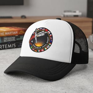 Puede incluir: Una gorra tipo trucker blanca y negra con un parche circular en la parte frontal. El parche muestra un gráfico de temática espacial con el texto "PROJECT HAIL MARY" y un anillo de banderas internacionales rodeando una nave espacial.