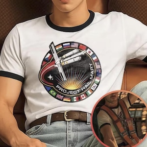 Puede incluir: Una camiseta blanca de estilo ringer con ribetes negros que presenta un gráfico circular de una nave espacial en el espacio, rodeada de banderas internacionales y el texto Project Hail Mary, con un destello de luz y una línea orbital roja.