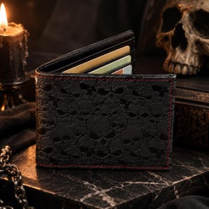 May include: A black bi-fold wallet featuring an embossed skull pattern across the exterior surface. The wallet is accented with red stitching along the edges and contains multiple slots holding several rectangular cards inside the top compartment.