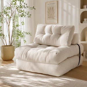 May include: A cream-colored, corduroy-textured floor chair featuring a tufted backrest and a thick, layered seat cushion. The chair is constructed from stacked, soft-touch fabric segments and includes a dark strap detail on the side.