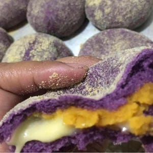 May include: A round purple bread roll with a light dusting of beige powder on the exterior. The roll is broken open to reveal a vibrant yellow filling and a creamy white, melted center, with several other similar rolls visible in the background.