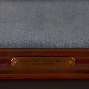 May include: A close-up of a wooden frame with a gold plaque that reads "Breaking the Moonlit Silence Frank C. McCarthy".