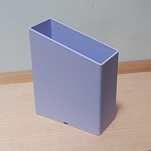 May include: A light lavender, rectangular storage container with a slanted top edge. The object features a tall, narrow profile with smooth, vertical walls and a hollow interior, constructed with a visible layered texture from 3D printing.