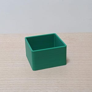May include: A small, square, open-top container with straight vertical walls. The object is made of a solid, matte green material and features a distinct layered texture on its exterior surfaces, consistent with a 3D printed construction.