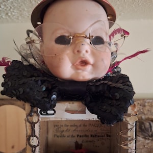 May include: A doll head wearing a red hat with a beetle ornament and clear glasses, adorned with a black sequined bow and feathers. The head sits atop a box featuring text that reads "pay to the order of the PACIFIC" and "Pacific National".