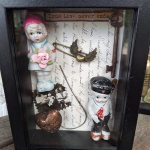 May include: A black shadow box frame containing two porcelain figurines, a metal key, a heart-shaped pin with wings, a copper heart, and a black and white photograph. The background features handwritten cursive text and the phrase True Love never ends.