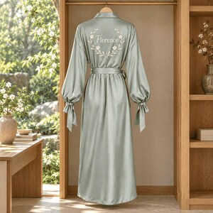 May include: A floor-length sage green satin robe featuring long sleeves with tie cuffs, a matching waist belt, and an embroidered floral wreath design on the back with the name Florence written in white script in the center of the wreath.