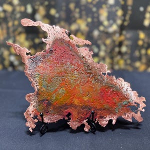 May include: An irregular, flat copper piece with a textured, oxidized surface featuring vibrant shades of red, orange, yellow, and green. The edges are jagged and raw, and the object is displayed on a small, black metal wire stand.