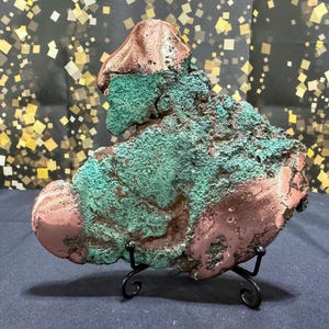May include: A raw mineral specimen featuring a combination of metallic copper-colored areas and a textured, vibrant green malachite surface. The irregular, organic-shaped rock is displayed upright on a black metal wire stand with curved feet.