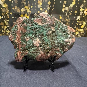 May include: A raw, irregular copper specimen featuring a textured surface with prominent metallic copper tones and oxidized green patina patches, displayed on a small, black, curved metal wire stand for stable upright positioning.