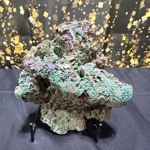May include: A raw mineral specimen featuring a porous, jagged texture with vibrant patches of teal, green, and deep purple coloration. The rock is displayed on a black metal stand, showcasing its irregular, craggy surface and natural crystalline structure.
