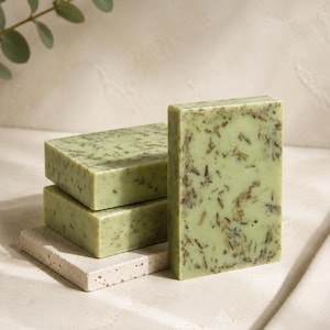 May include: Three rectangular bars of light green soap with visible dark botanical inclusions. Two bars are stacked horizontally, while the third stands vertically in the foreground, showcasing a smooth texture with scattered dried herbal pieces.