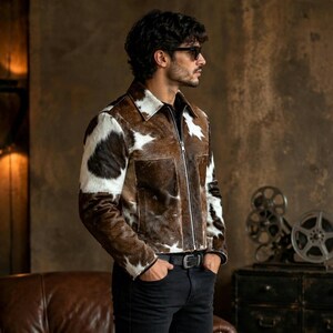 May include: A long-sleeve jacket featuring a brown and white cowhide pattern with a front zipper closure, a pointed collar, and two large rectangular front pockets. The jacket is worn with black pants featuring a black belt with a silver buckle.