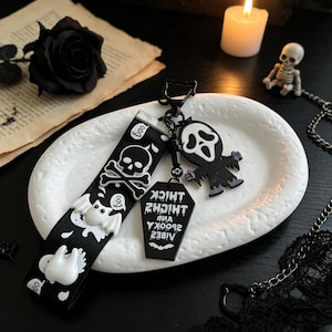 May include: A black fabric keychain strap featuring white printed skulls, bats, and the word "boo," attached to a black metal clasp. It includes a black coffin-shaped charm with the white text "THICK THIGHS AND SPOOKY VIBES" and a ghost-like figure charm.