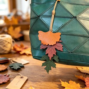 May include: A dark green leather handbag with a geometric paneled design features a hanging charm made of three layered leather leaves in orange, reddish-brown, and dark green, suspended by a thin light tan leather cord from the top handle.