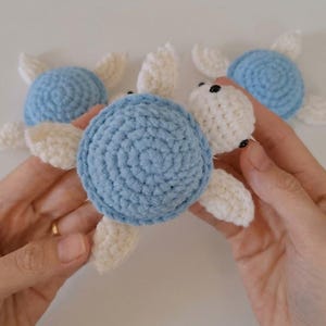 May include: Three small crocheted sea turtles featuring light blue circular shells and off-white heads and flippers. Each turtle has two small black bead eyes. The yarn texture is visible throughout the handmade figures.