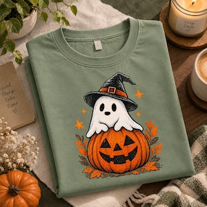 Könnte beinhalten: Ein salbeigrünes T-Shirt mit Rundhalsausschnitt und einem Grafikdruck eines weißen Geistes mit schwarzem Hexenhut, der auf einem geschnitzten orangefarbenen Kürbis sitzt, umgeben von orangefarbenen Blättern und kleinen Sternen.