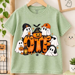 May include: A sage green short-sleeve t-shirt featuring a graphic of several white ghosts and a central jack-o'-lantern wearing a witch hat. The design includes the orange text "OTF" with spiderweb accents, a lantern, and small bats.