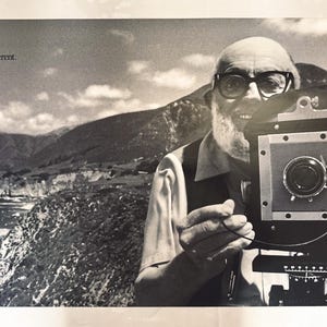 May include: A black and white poster featuring a portrait of a bearded person with glasses operating a large format camera. The text Think different. and a small rainbow-colored apple logo appear in the top left corner above a coastal landscape.