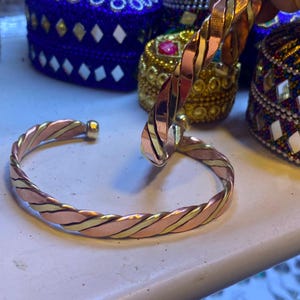 Peut inclure: Un bracelet manchette au design torsadé composé de brins alternés en cuivre poli et en laiton. Le bracelet présente une forme circulaire ouverte avec des extrémités arrondies et lisses, mettant en valeur une finition métallique.