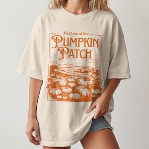May include: An off-white, short-sleeved t-shirt featuring a large, orange graphic print. The design includes the text 'Meet me at the PUMPKIN PATCH' above an illustration of a field filled with pumpkins and rolling hills in the background.