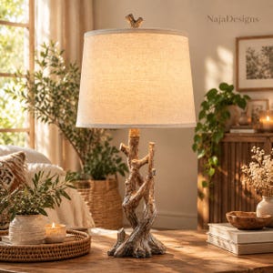 May include: A table lamp featuring a base sculpted to resemble intertwined tree branches with a textured, weathered brown finish. The lamp is topped with a tapered, off-white fabric drum shade and a small branch-shaped finial at the top.