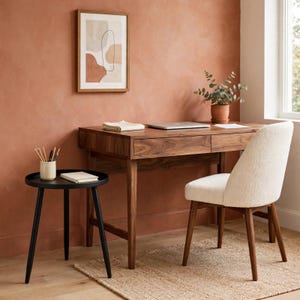 May include: A dark wood desk with two drawers, a cream-colored upholstered chair with wooden legs, and a small black round side table. On the desk sits a laptop and a folded cloth. The side table holds a cup with pencils and a small book.